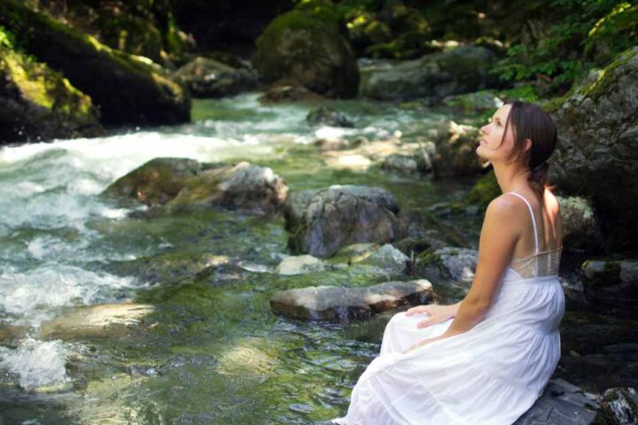 A woman sitting by a stream in the forest, deep in contemplation.