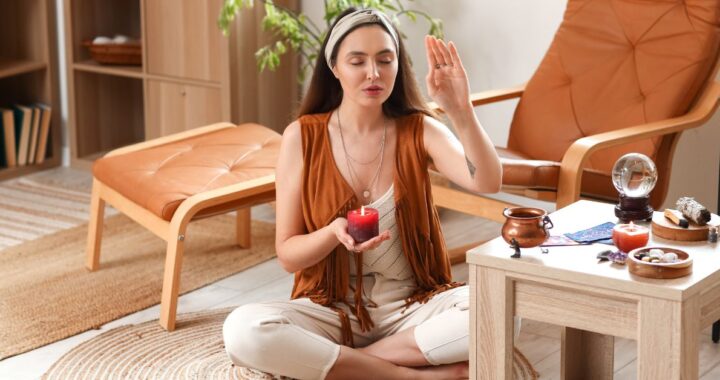 A woman meditating with closed eyes, holding a red candle in one hand and making a mystical hand gesture (mudra) in a bright, private room filled with tarot cards, crystals, and candles.