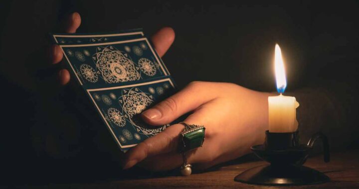 Female hands shuffling a deck of tarot cards in the dark with a flickering candle nearby.