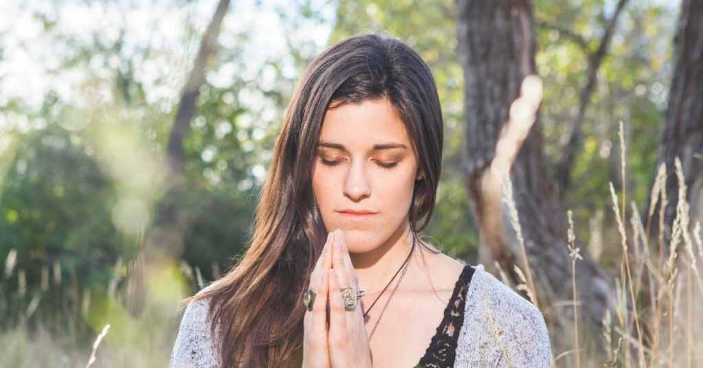 A woman with closed eyes, hands clasped in prayer, stands peacefully in a sunlit forest clearing. This image is meant to demonstrate the 3rd of 6 spiritual practices for improving your intuition.