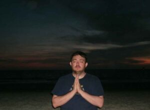 John Culbertson sits in meditation with eyes closed on a quiet beach at sunset, hands in prayer position. For the blog post what happens during a session with John Culbertson